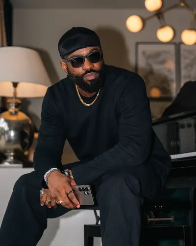 Brawks leans on a grand piano in shades and gold chain, phone in hand under soft ambient light