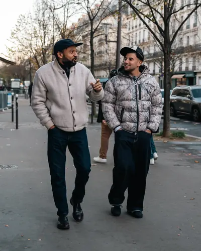 Riadh and Fabrice Éboué stroll side by side down a tree-lined Parisian boulevard, deep in conversation under the autumn light.